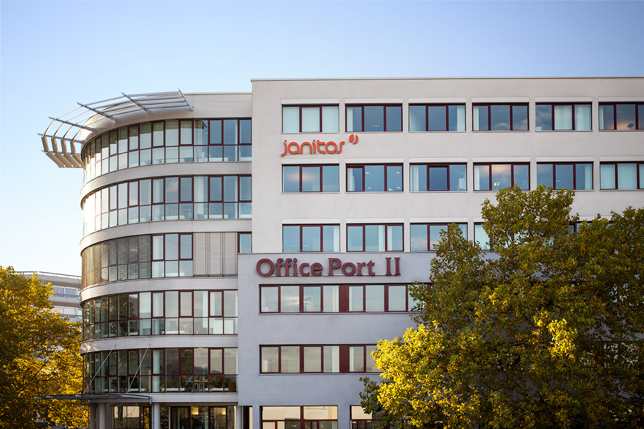 Die Außenansicht des Janitos-Firmengebäudes in Heidelberg mit einer großen, ums Eck gebogenen Glasfassade - dem orangenen Janitos Logo und einer sanften Morgensonne, die sich im Glas spiegelt